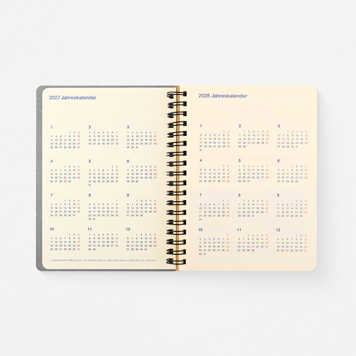 An open rollbahn spiral-bound planner or calendar showing the interior pages for 2027 and 2028, with small monthly grids printed in muted blue, red on a warm cream page,